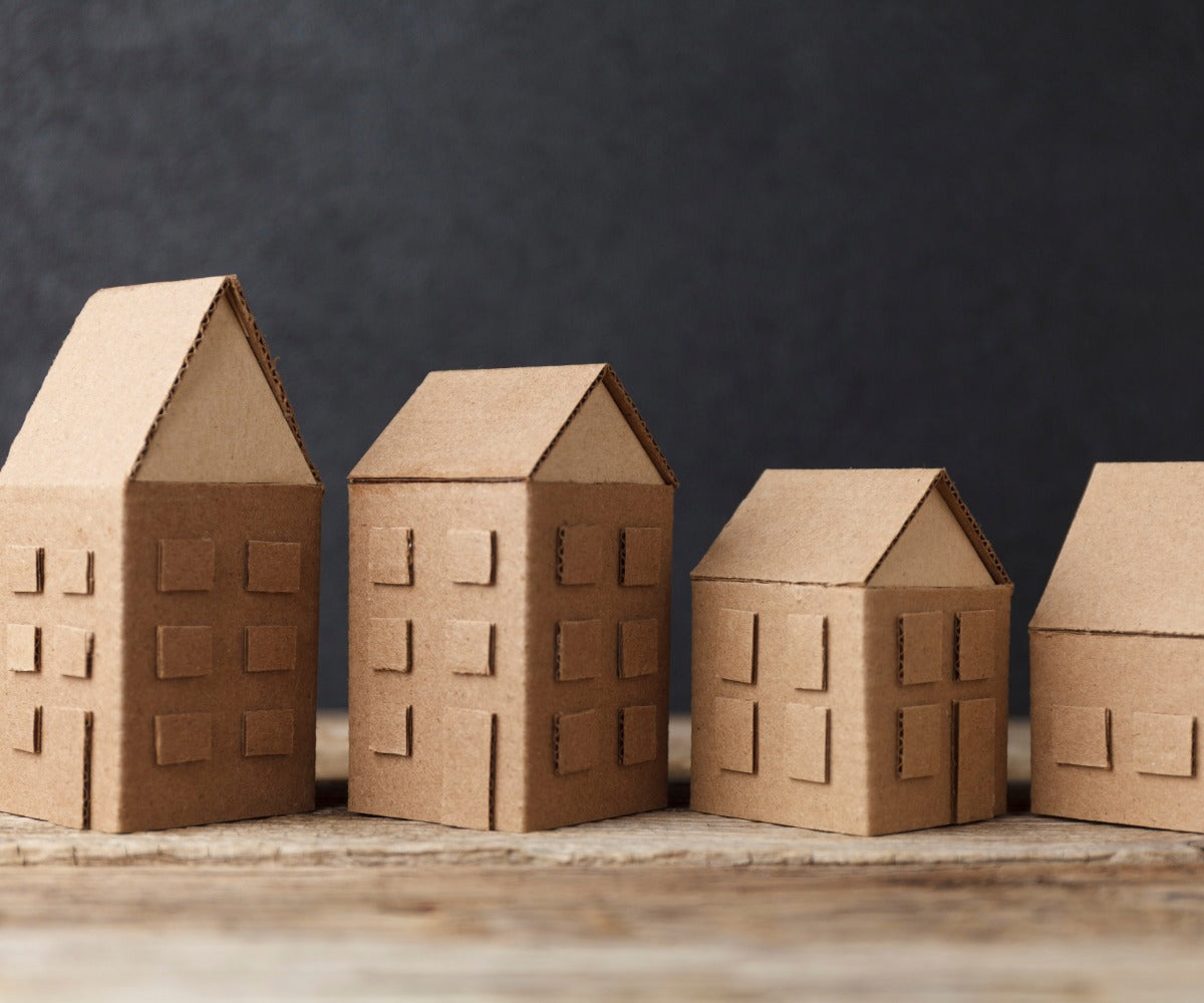 Four cardboard house models arranged in a row on a wooden surface with a dark background.